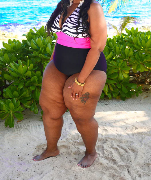 Woman in a black and pink swimsuit standing on a sandy beach with ocean and palm trees in the background.