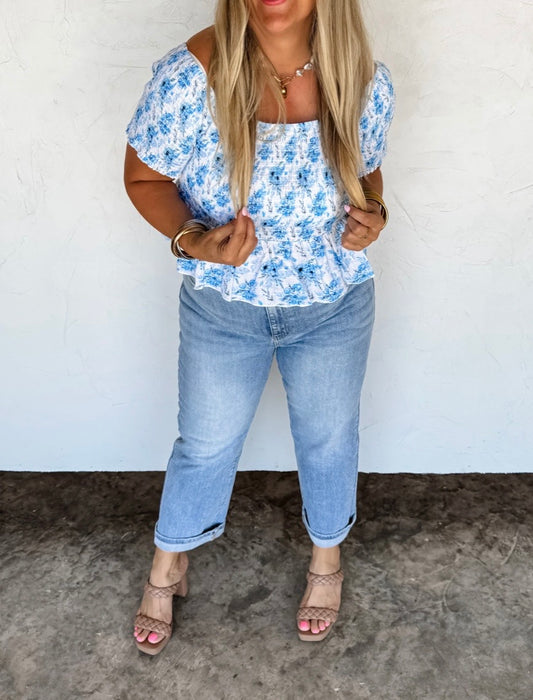 Woman wearing a blue floral top and jeans against a white wall.