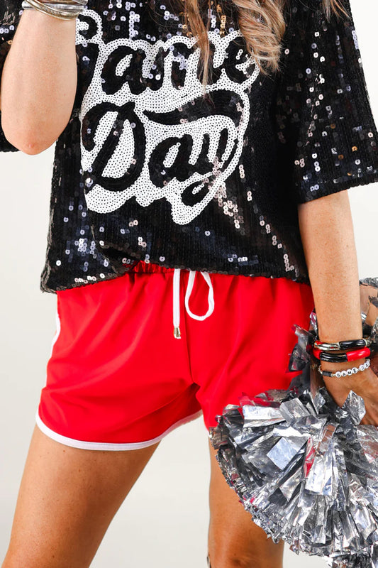 Person wearing a black sequin top with 'Game Day' text, red shorts, and holding silver pom-poms.