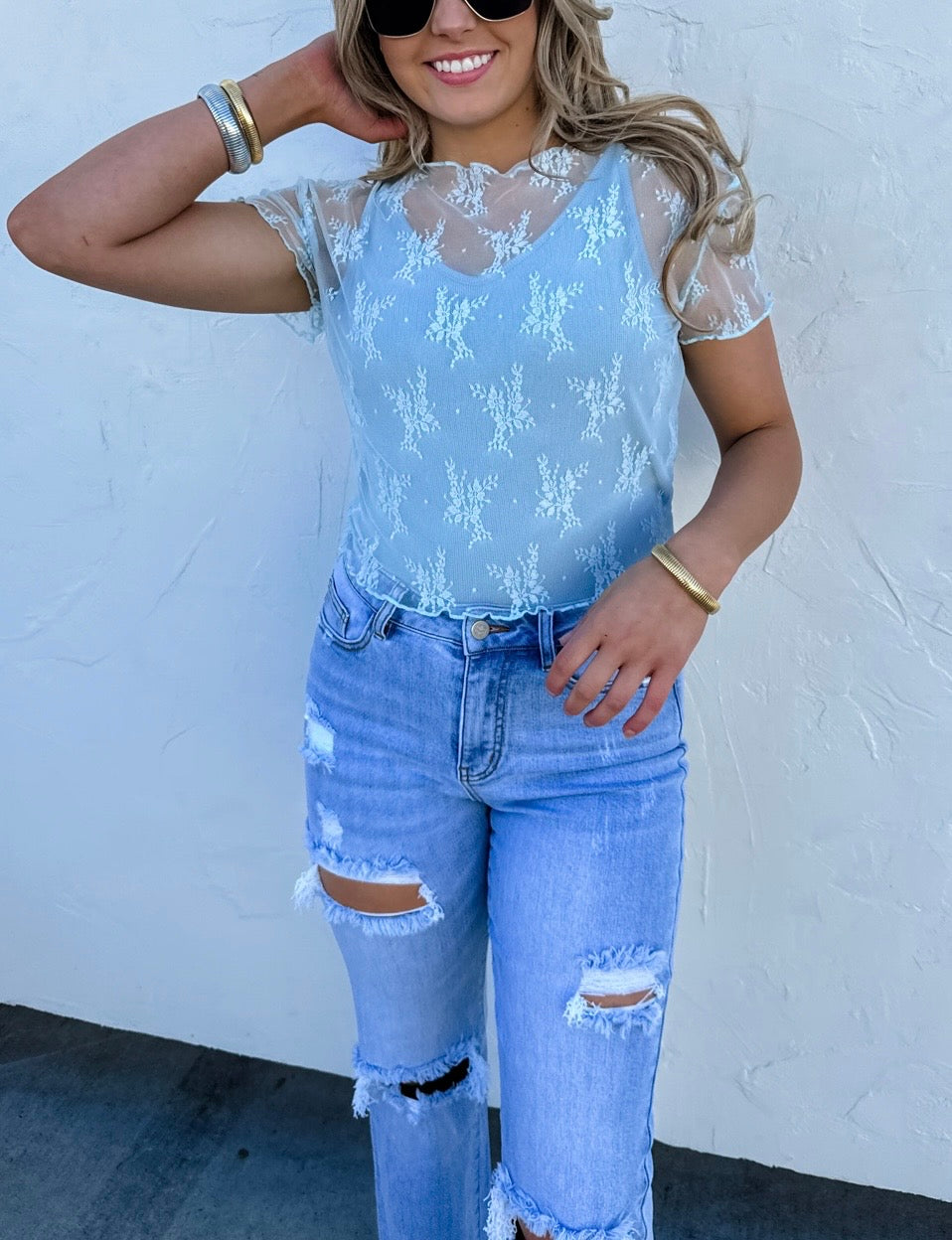 Woman wearing a light blue lace top and ripped jeans against a white wall.