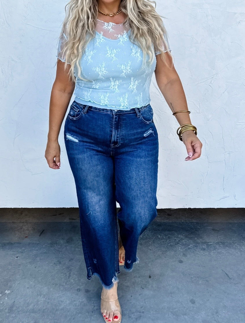 Person wearing a light blue lace top and blue jeans against a white wall.