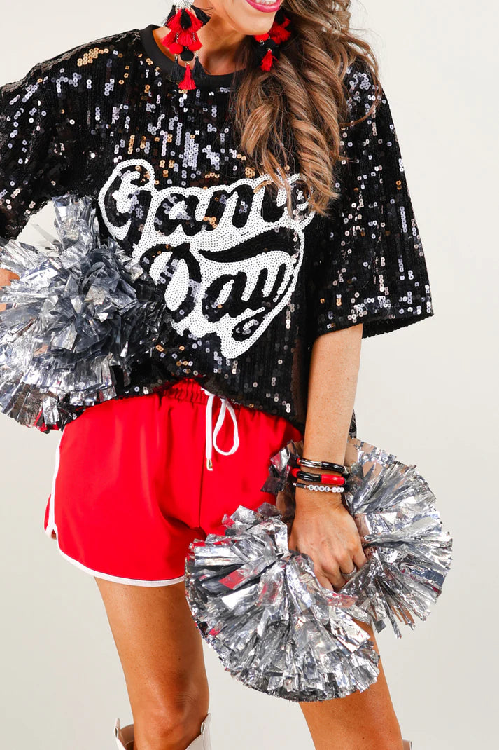 Person wearing a black sequin 'Game Day' shirt with red shorts, holding silver pom-poms.