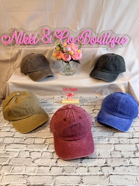 Four baseball caps in different colors on a table with 'Nikki & Lo Boutique' sign.