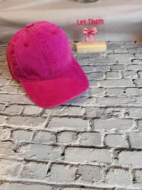 Pink cap on a textured surface with 'Let Them' text and bow in the background